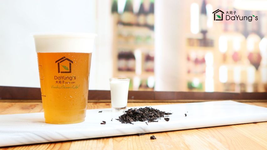 Plastic cup of iced tea with logo beside a small white cup and loose tea leaves on a wooden table.
