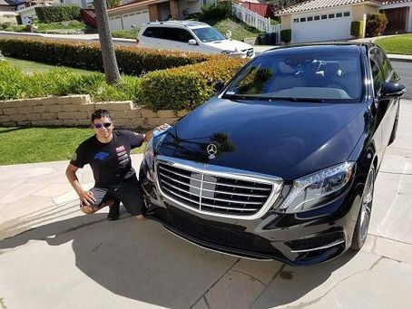 Man posing next to a black Mercedes-Benz sedan in a driveway on a sunny day.