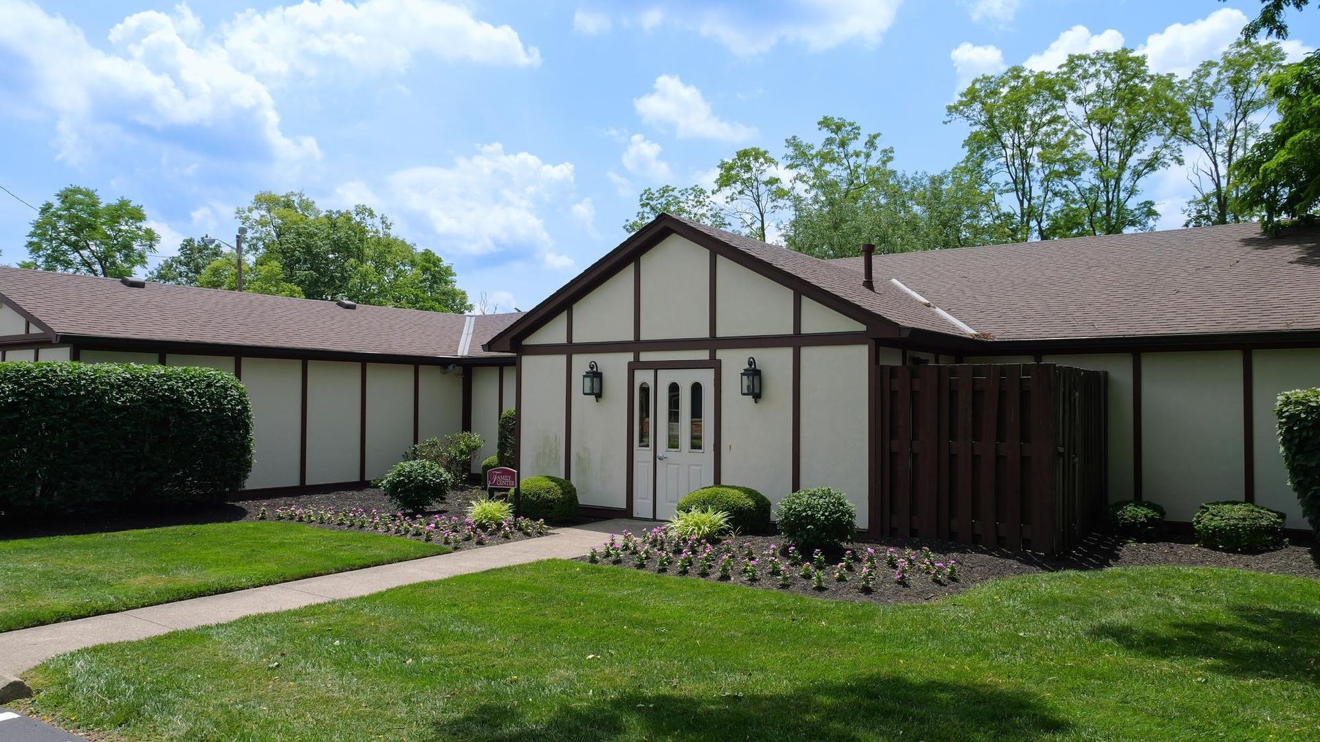 Single-story beige house with brown roof, front lawn, and trees under a blue sky