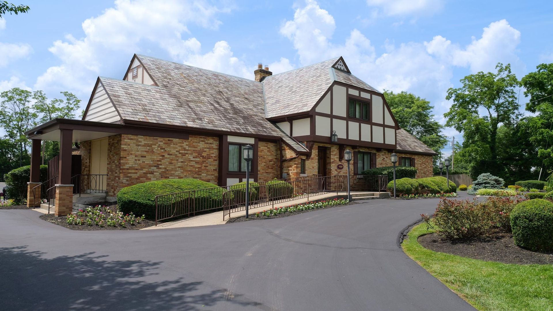 Tudor-style brick building with a shingled roof and landscaped driveway under a blue sky