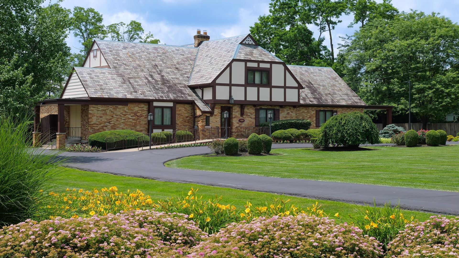 Stone house with a gabled roof, front lawn, and curved driveway amid trees and flowers