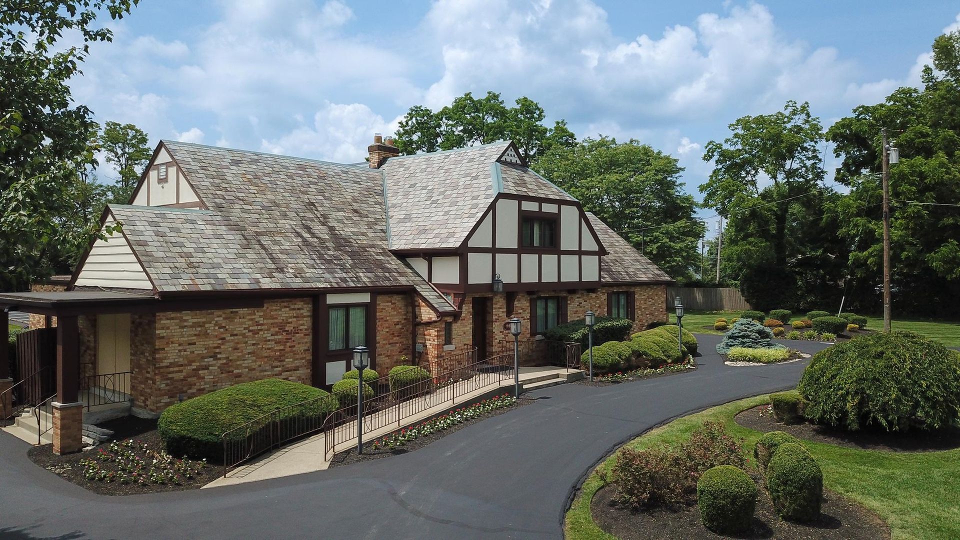 Large Tudor-style house with a brick entrance, curved driveway, and landscaped trees under a blue sky.