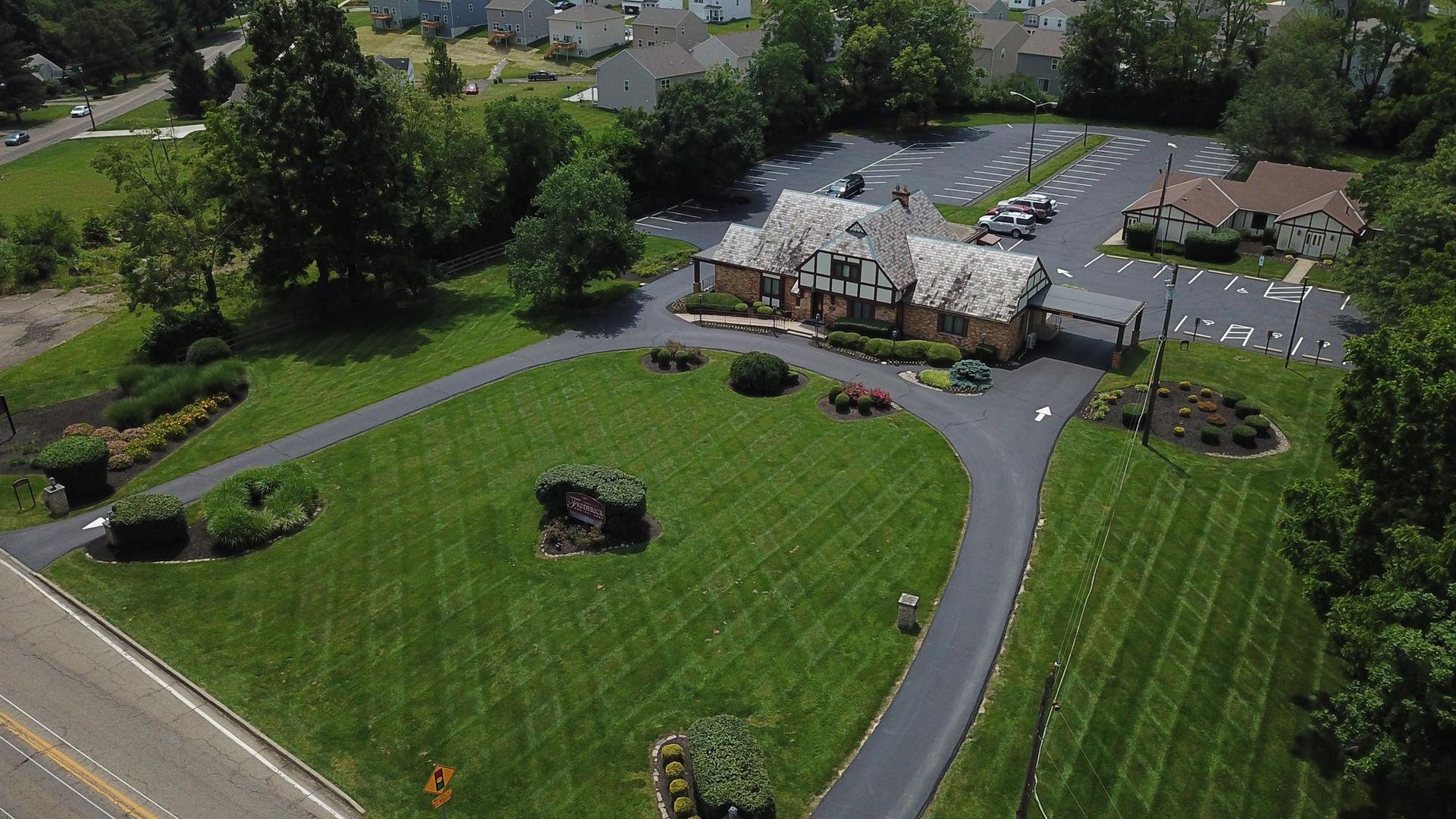Aerial view of a white building beside a parking lot and winding driveway, surrounded by green lawns and trees