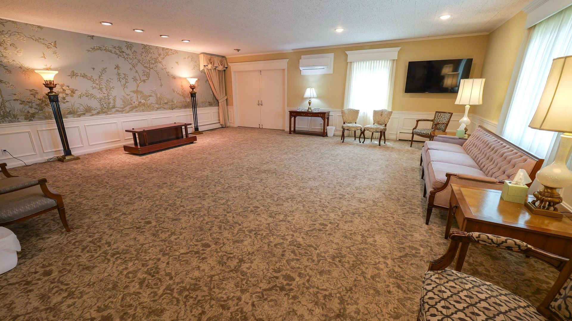 Spacious beige living room with carpet, sofa, wall-mounted TV, table lamps, and decorative wall art