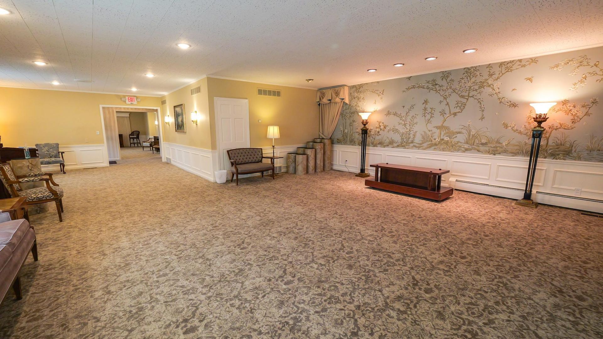 Spacious beige lounge with patterned carpet, benches, and wall sconces along a long hallway
