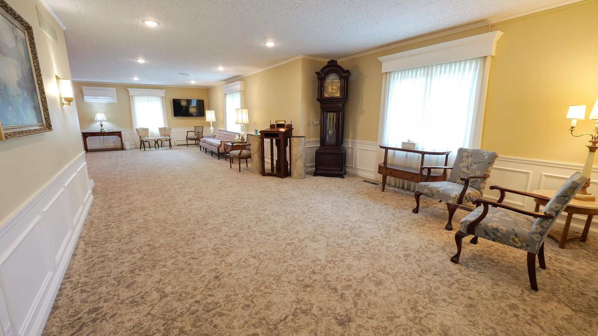 Spacious beige-carpeted lobby with armchairs, tables, lamps, and a grandfather clock by bright windows