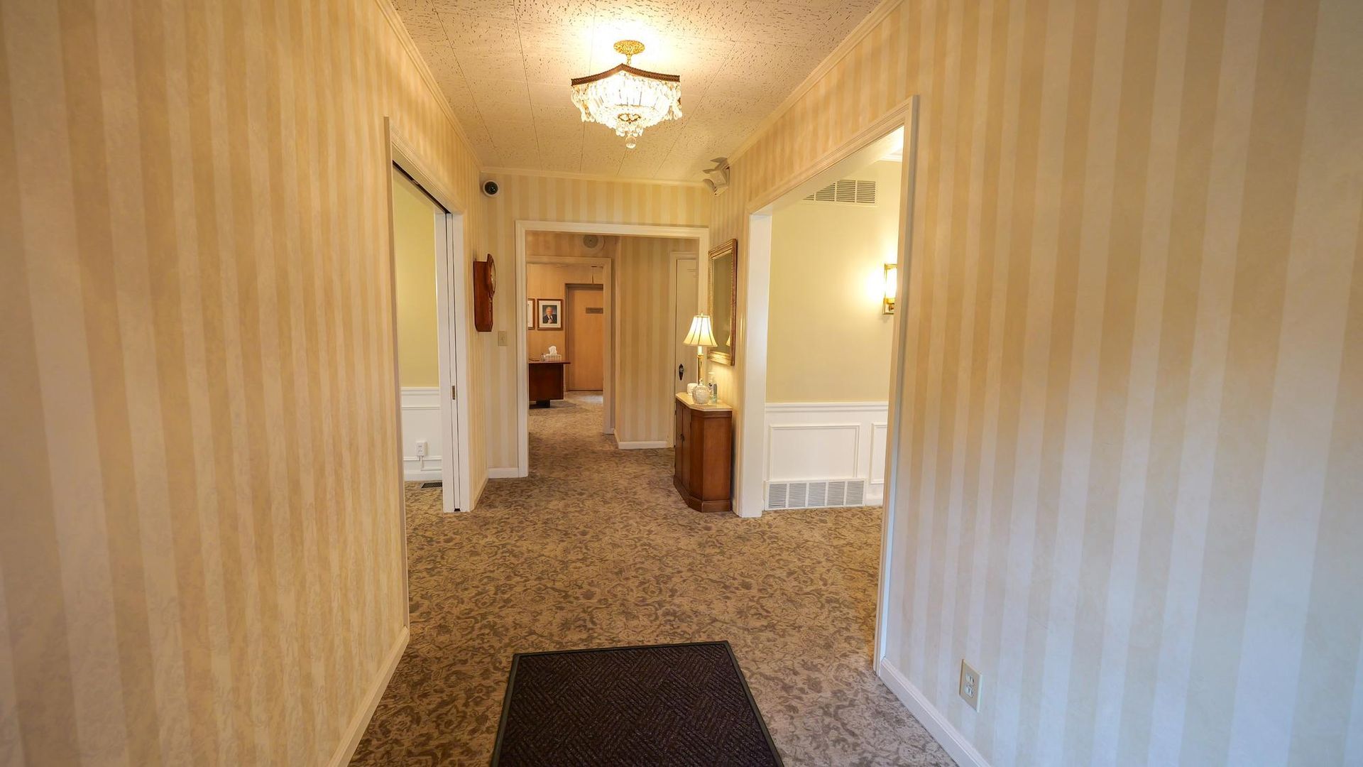 Long carpeted hallway with striped wallpaper, ceiling lights, and a small table lamp at the far end