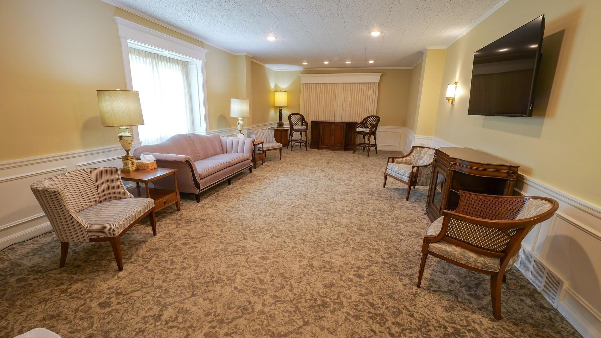 Spacious carpeted lounge with sofas, chairs, lamps, and a wall-mounted TV.