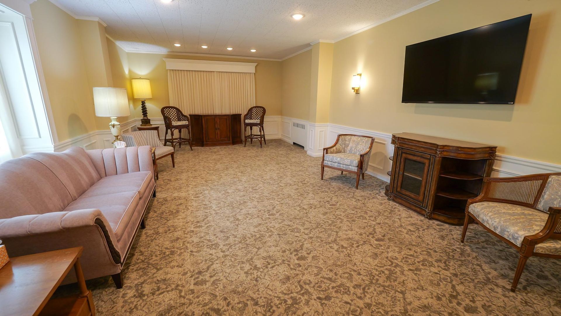 Spacious living room with beige carpet, two sofas, chairs, lamps, and a wall-mounted TV.