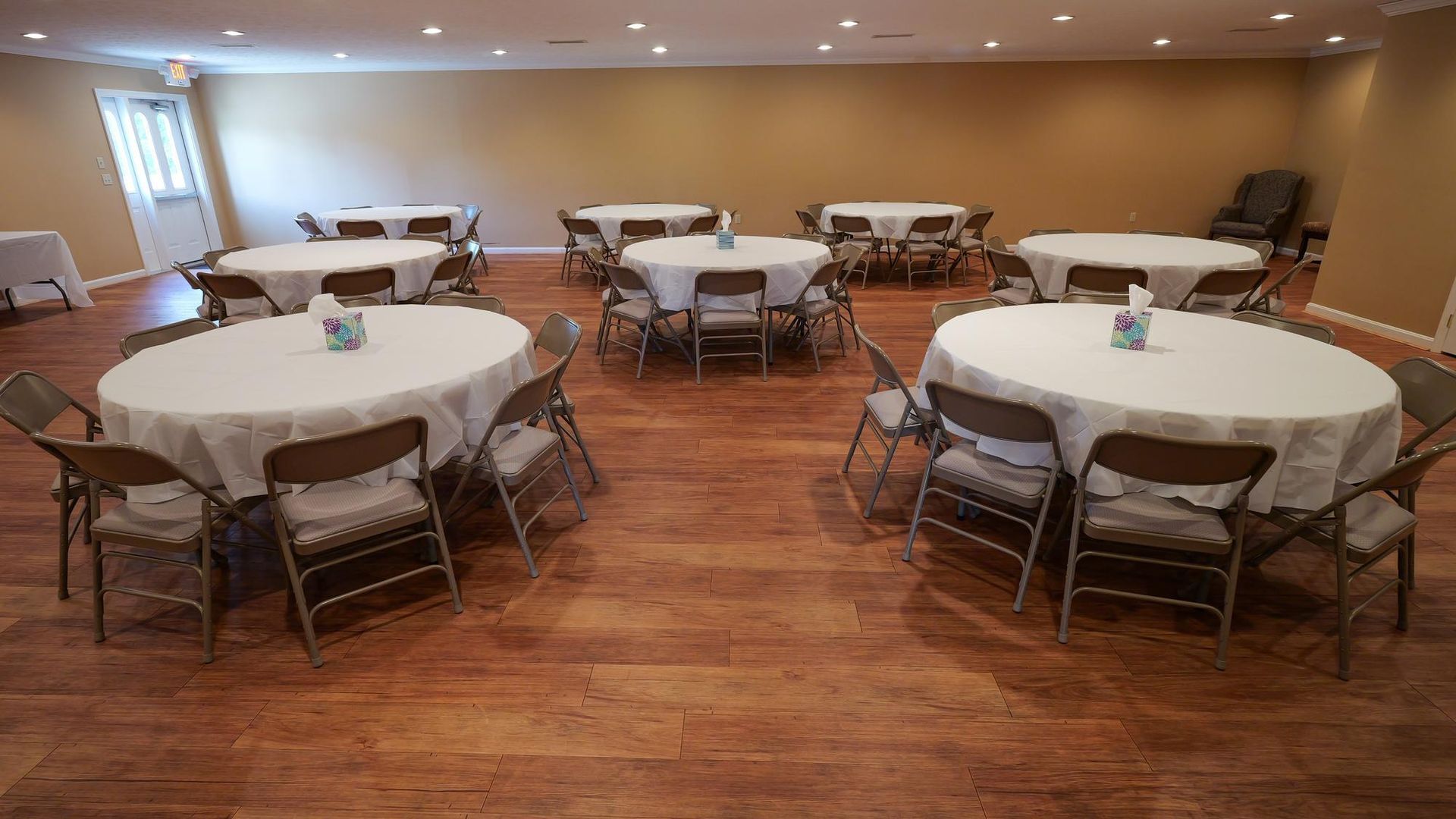 Empty banquet room with round tables, folded chairs, and wood flooring under warm ceiling lights