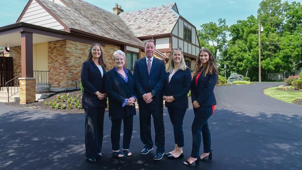 Five people in business attire standing on a driveway outside a building, smiling at the camera.