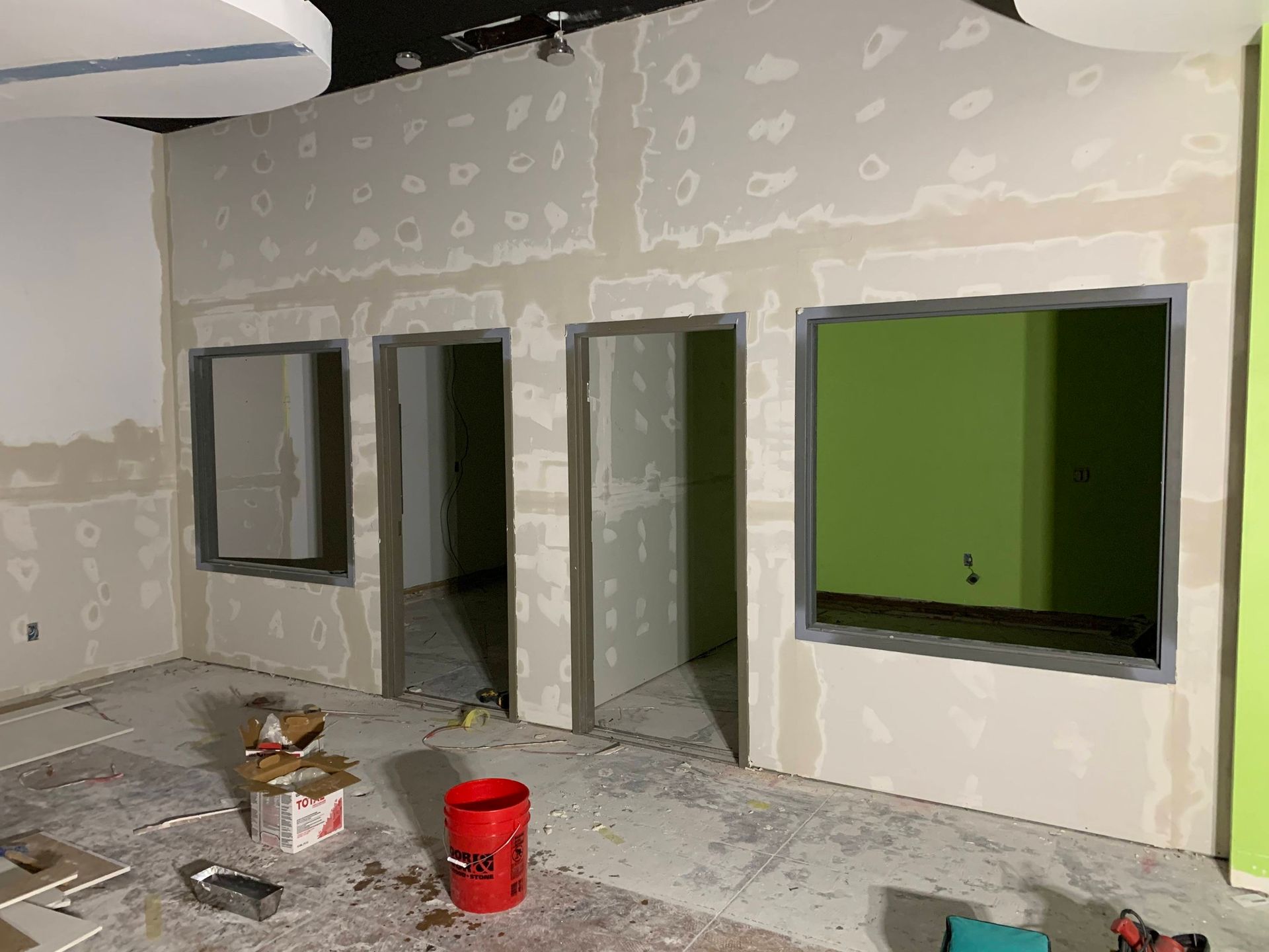 Indoor room under renovation with unfinished drywall, window openings, and paint buckets on the floor
