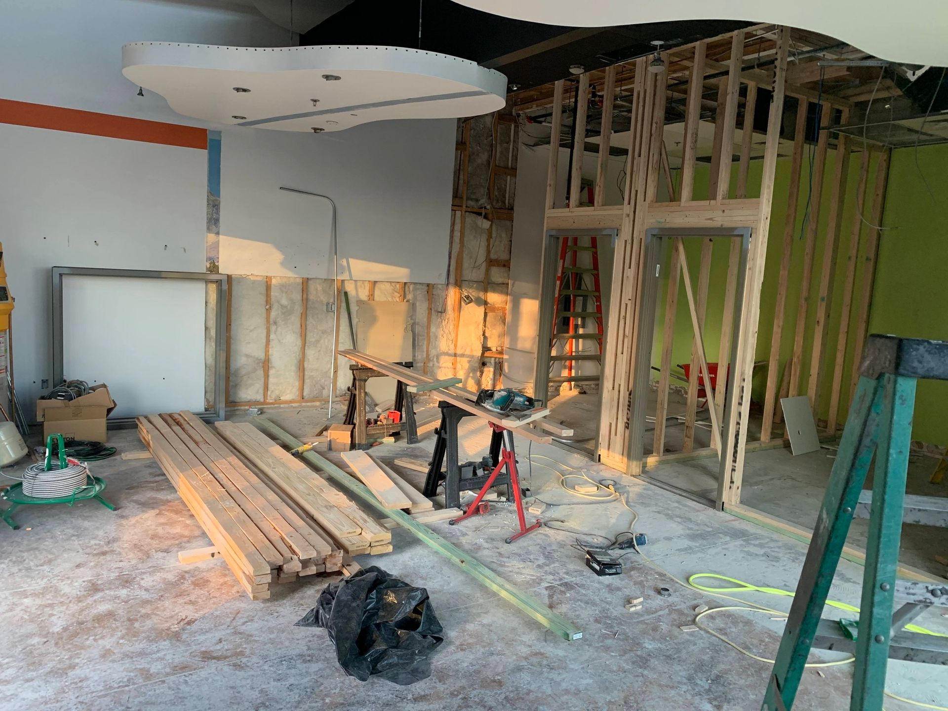Renovation site with exposed framing, stacked lumber, tools, and dust-covered floor inside a room