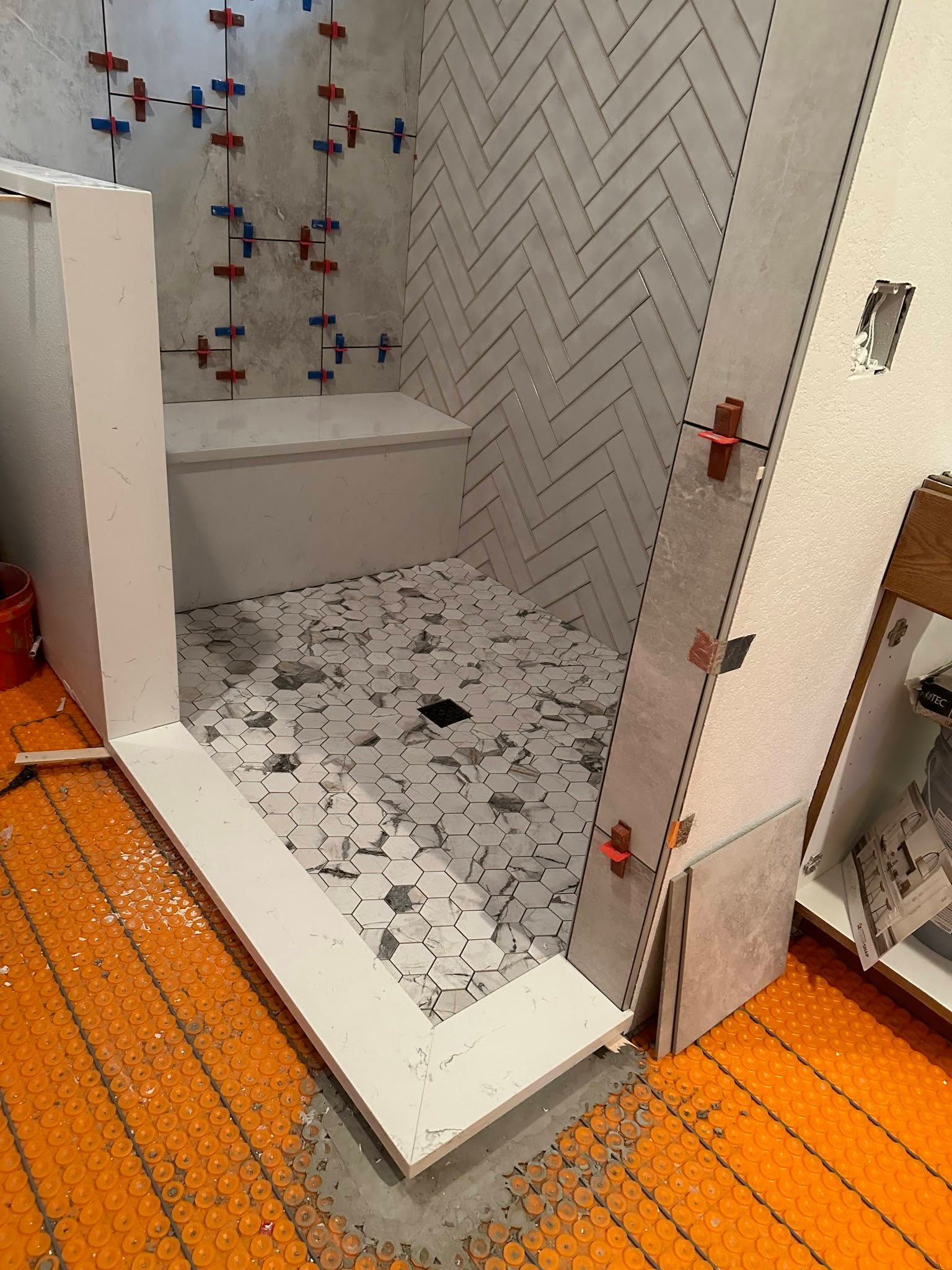 Small tiled bathroom shower with orange floor and white patterned wall tiles, viewed through open door.