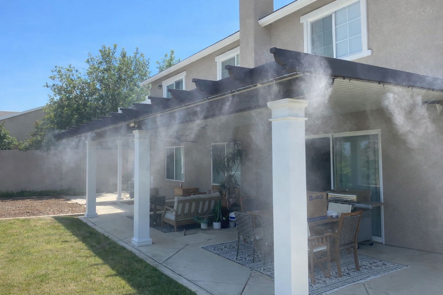 Backyard patio with white columns and chairs, spraying water from a misting system under a sunny sky