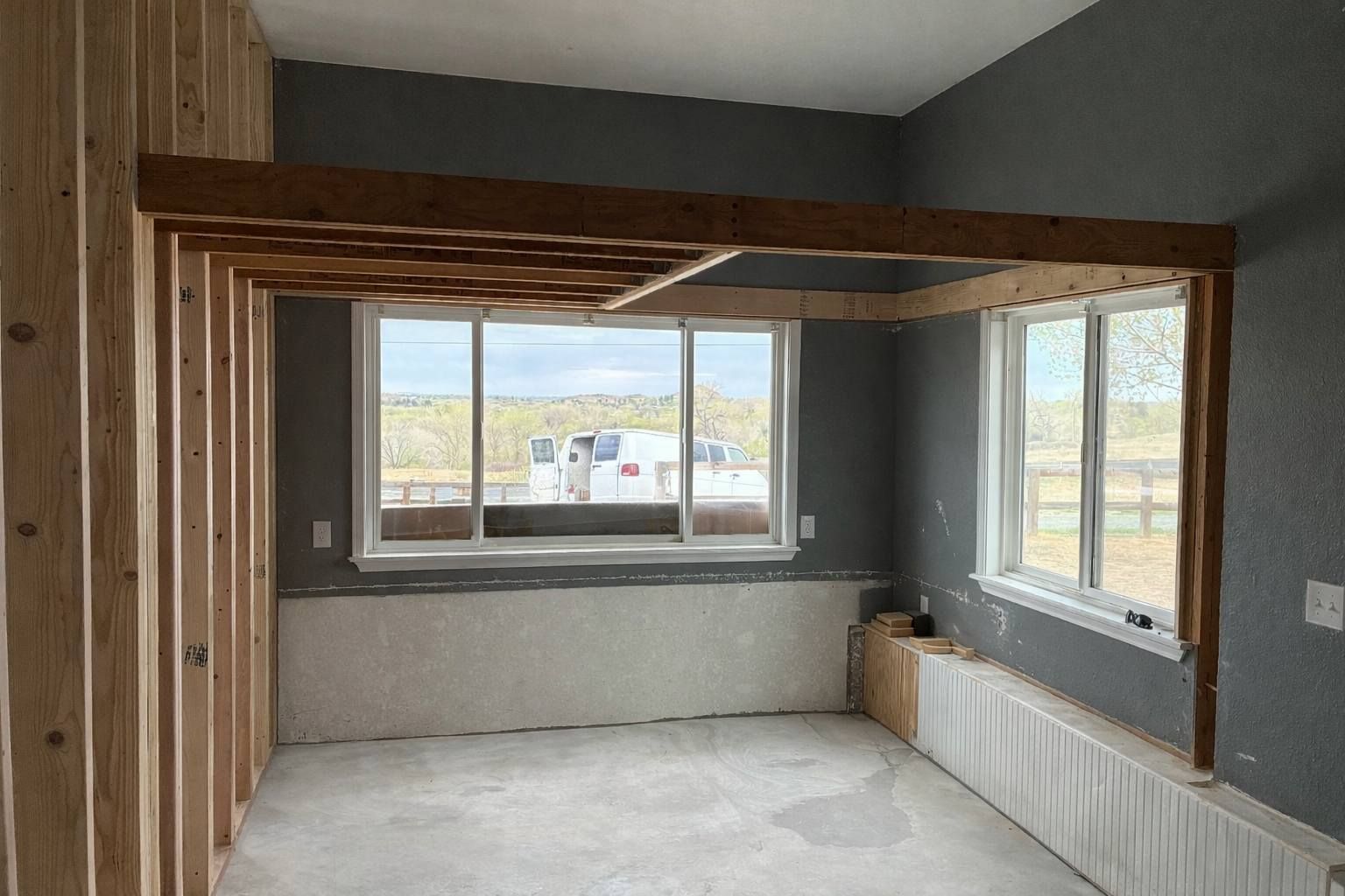 Empty gray room with exposed wood framing and large windows overlooking a landscape