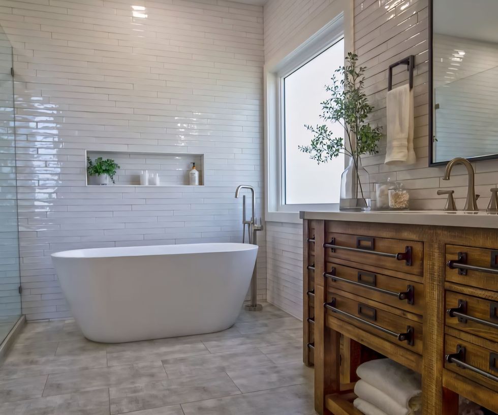 Bright bathroom with freestanding tub, white tile shower, window, and wooden vanity with marble top