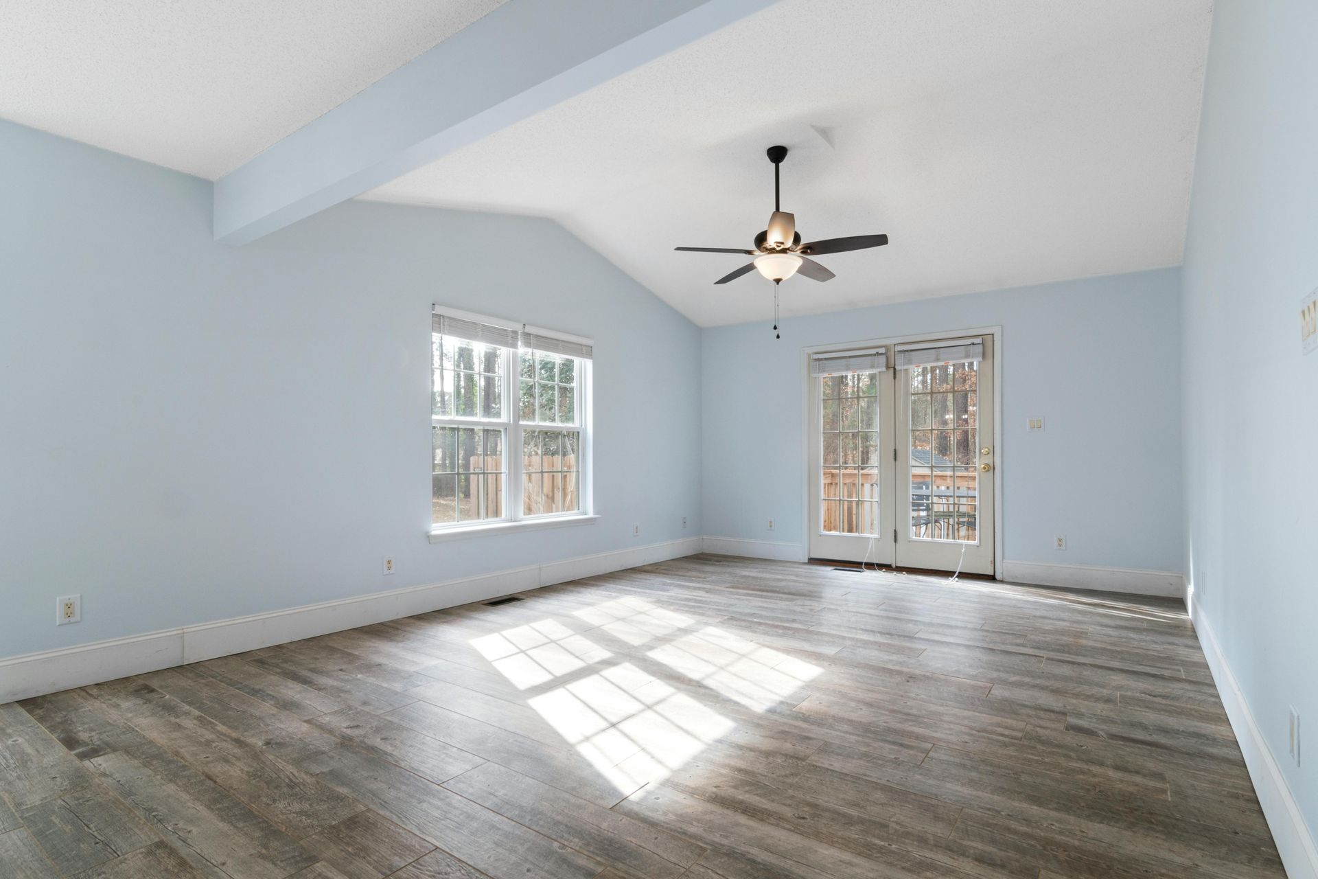 Empty light-blue living room with vaulted ceiling, ceiling fan, windows, and sunlit floor.
