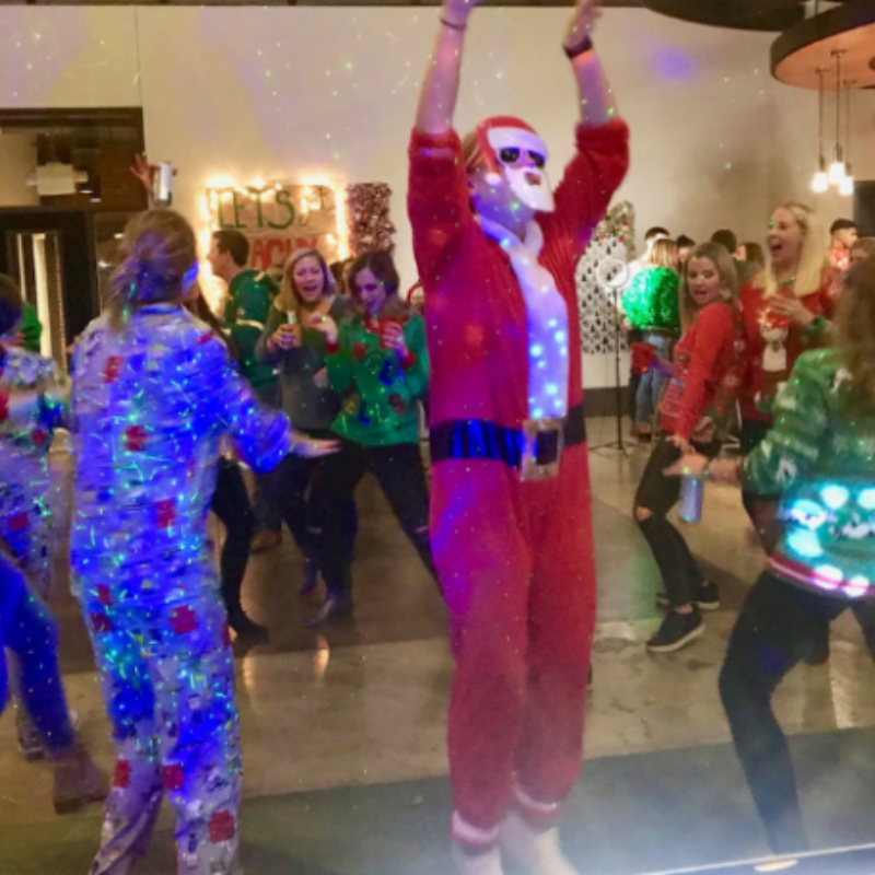 A person in a Santa costume with sunglasses jumps in a room filled with people wearing festive holiday sweaters.