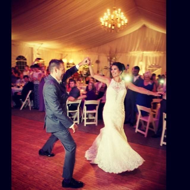 A couple dances on a wooden floor in a tented wedding reception, the person in the white gown twirling as they hold hands.