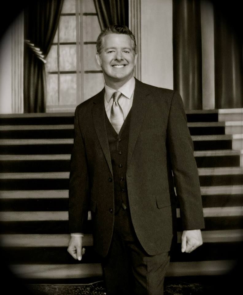 A smiling person in a formal suit and vest standing on a staircase in front of a window with draped curtains.