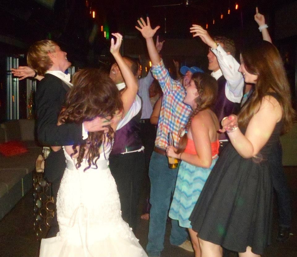 A wedding party dancing at an indoor reception, with guests in formal attire and colorful clothing raising their hands.