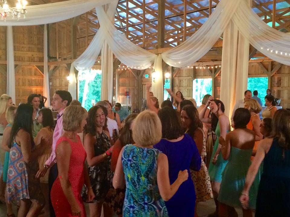 People dance in a rustic wooden event venue decorated with white drapery and hanging lights.