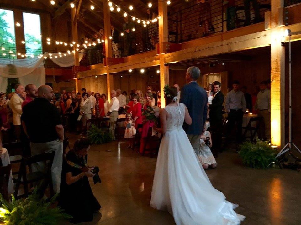 A bride in a white wedding gown walks down the aisle of a rustic wooden barn venue filled with guests under string lights.