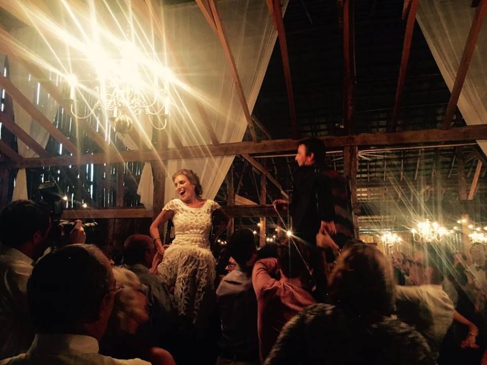 A bride and groom are lifted into the air by wedding guests inside a rustic barn illuminated by hanging chandeliers.