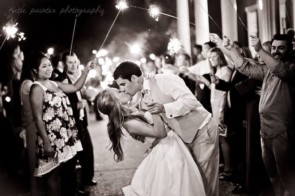 A bride and groom in wedding attire kiss as they dip while guests hold sparklers at night.
