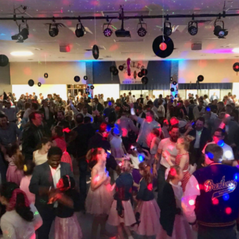 A crowded dance floor at a themed party with colorful disco lights, decorations hanging from the ceiling, and many people.