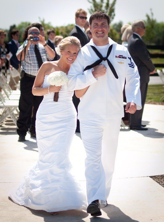 A bride and a man in a US Navy dress white uniform walking arm-in-arm outdoors at a wedding.