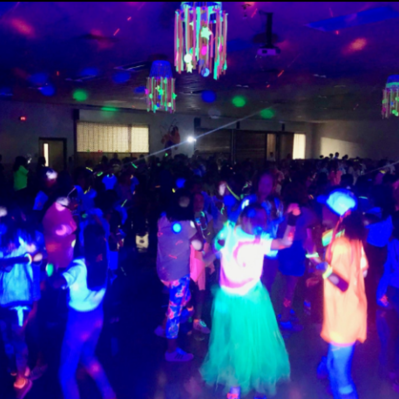 A crowd of people wearing neon clothing dances in a dimly lit, black-lit room decorated with glowing hanging streamers.