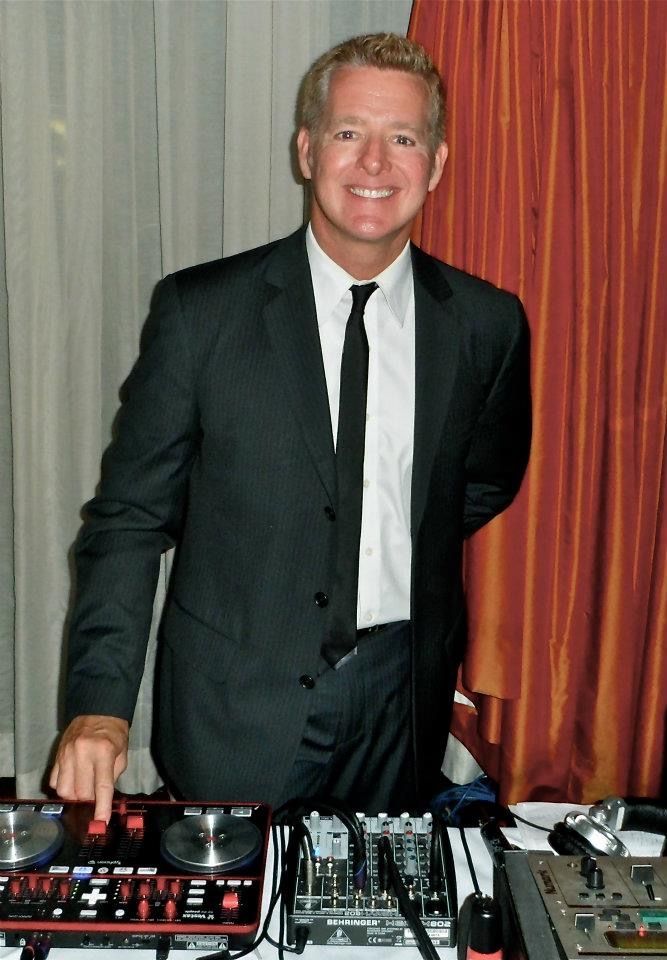 A smiling person in a suit stands behind a DJ controller and mixer in front of draped curtains.