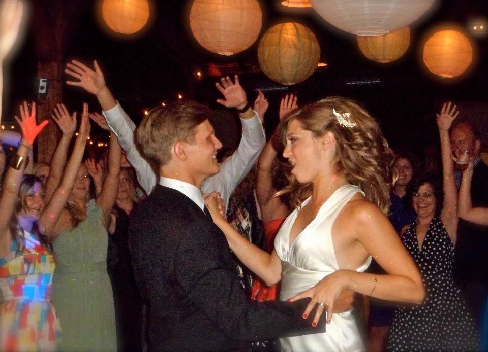 A couple dances at a dimly lit indoor event with paper lanterns, surrounded by guests with their hands raised.