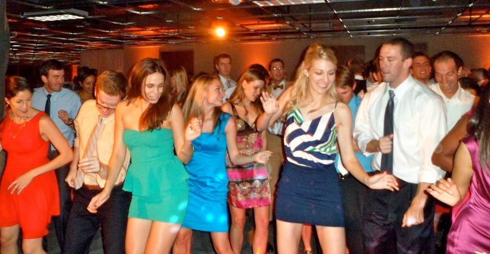A group of people dance together at an indoor formal event, wearing vibrant dresses and dress shirts.
