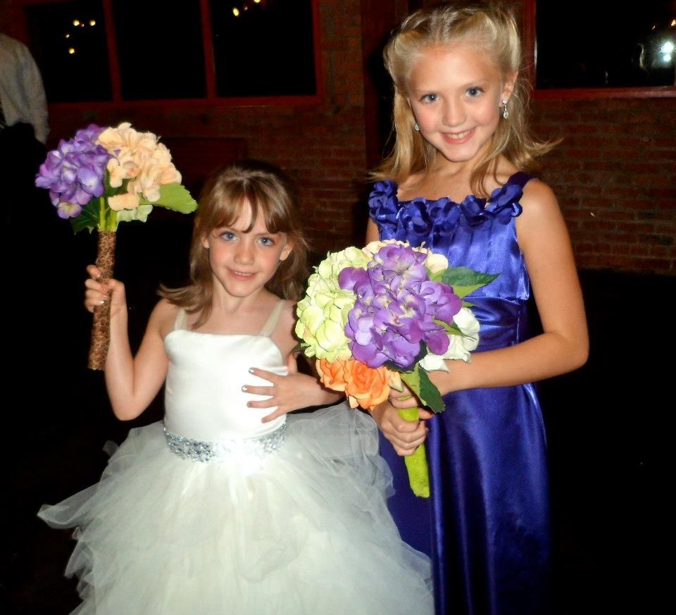Two children in formal attire hold flower bouquets while posing indoors against a brick wall.