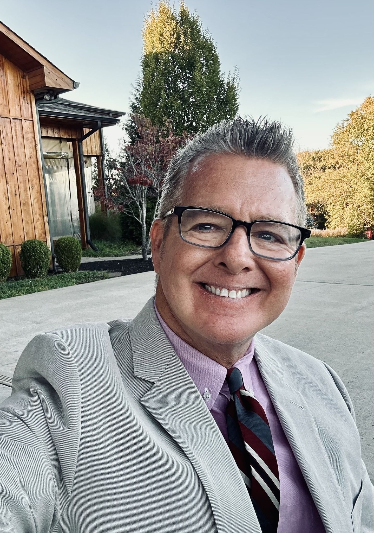 Smiling person in a gray suit and tie taking a selfie outdoors near a building and trees
