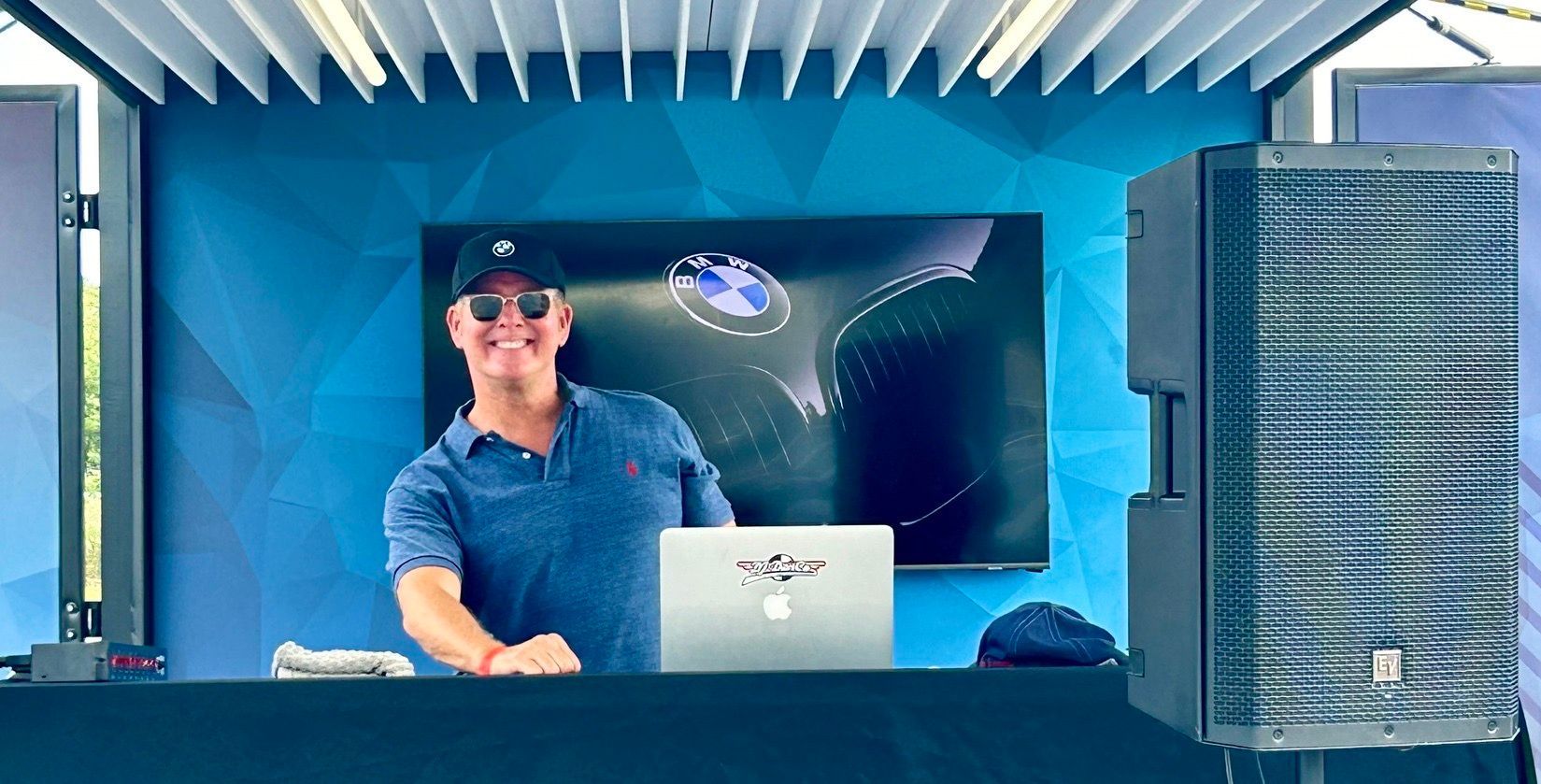 A smiling person wearing a cap and sunglasses stands behind a DJ table with a laptop in a blue, geometric-patterned booth.