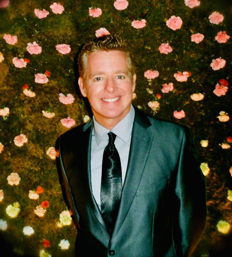 A smiling person in a dark suit and tie, posed in front of a wall decorated with pink flowers.