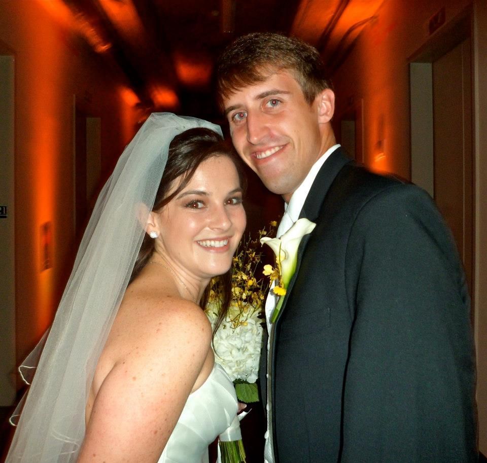 A smiling couple in wedding attire poses together in a dimly lit, orange-toned hallway.