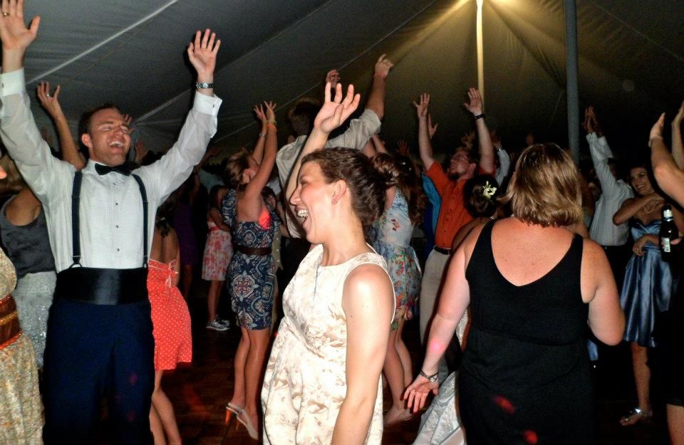 People dance with hands raised in a tented venue, including a man in a tuxedo and a woman in a light dress.
