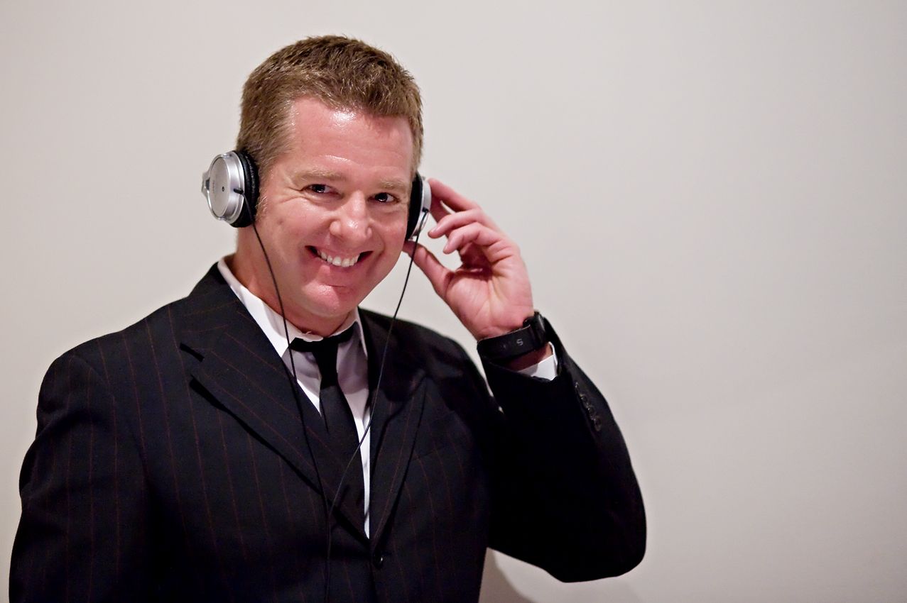 A smiling person wearing a black suit, white shirt, and black tie, holding a pair of headphones over their ears.