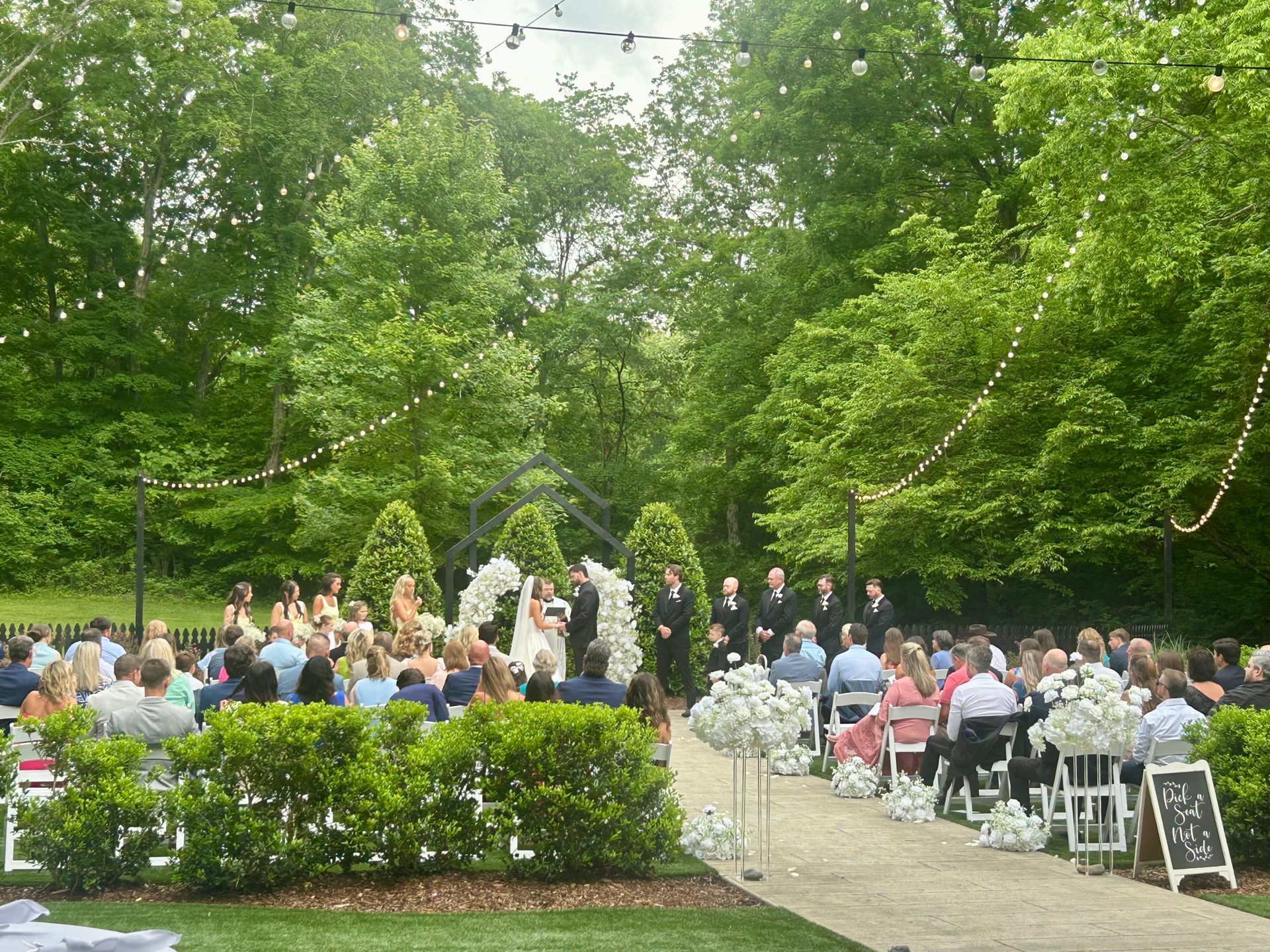 Outdoor wedding ceremony with guests seated in a garden, facing the couple under trees and white chairs