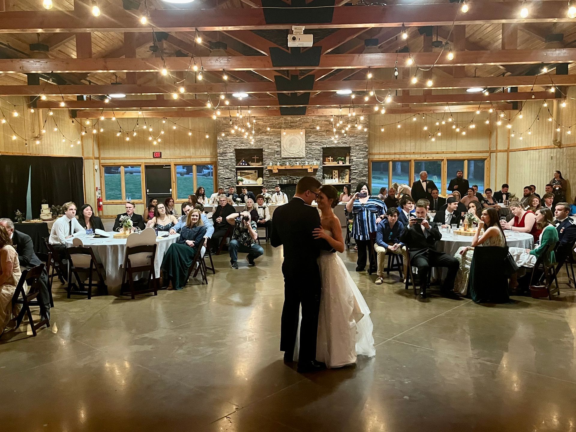 Couple slow dancing at a reception in a warmly lit hall with seated guests סביב tables