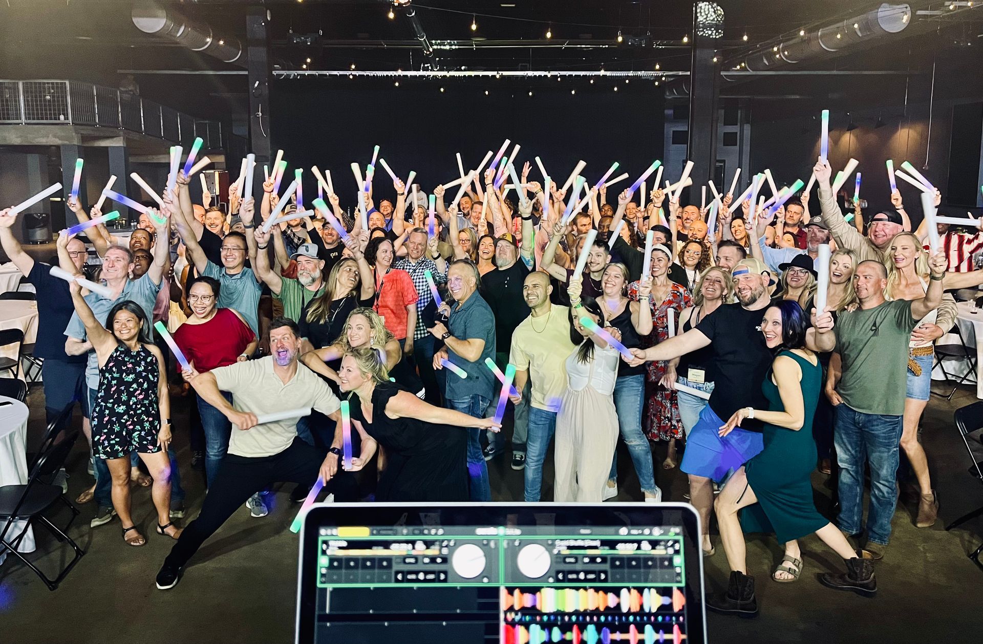 Large group dancing and cheering in a dim concert venue, with stage lights and DJ equipment in the foreground