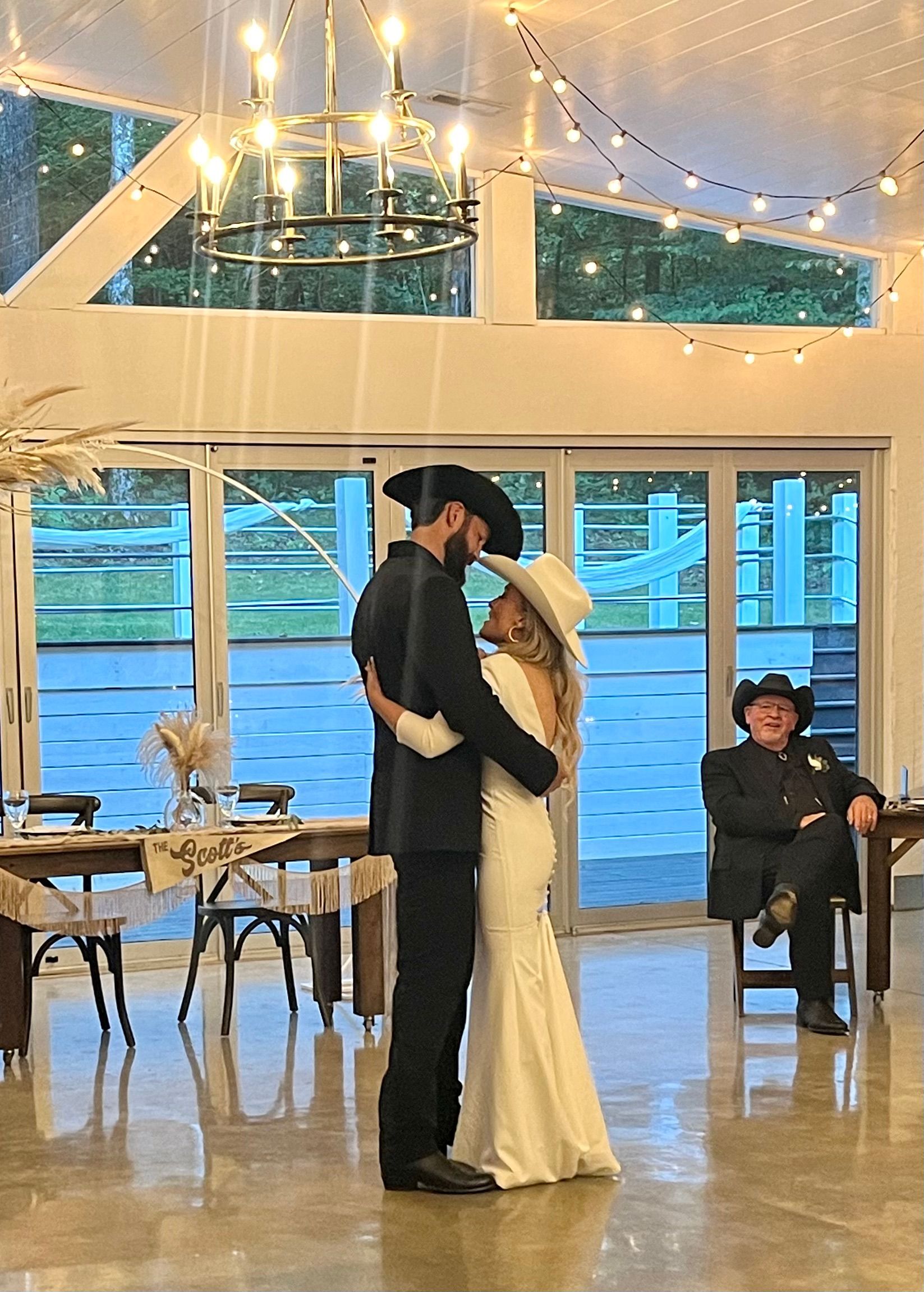 Couple in black and white embrace at a wedding reception under string lights, with a seated guest nearby.