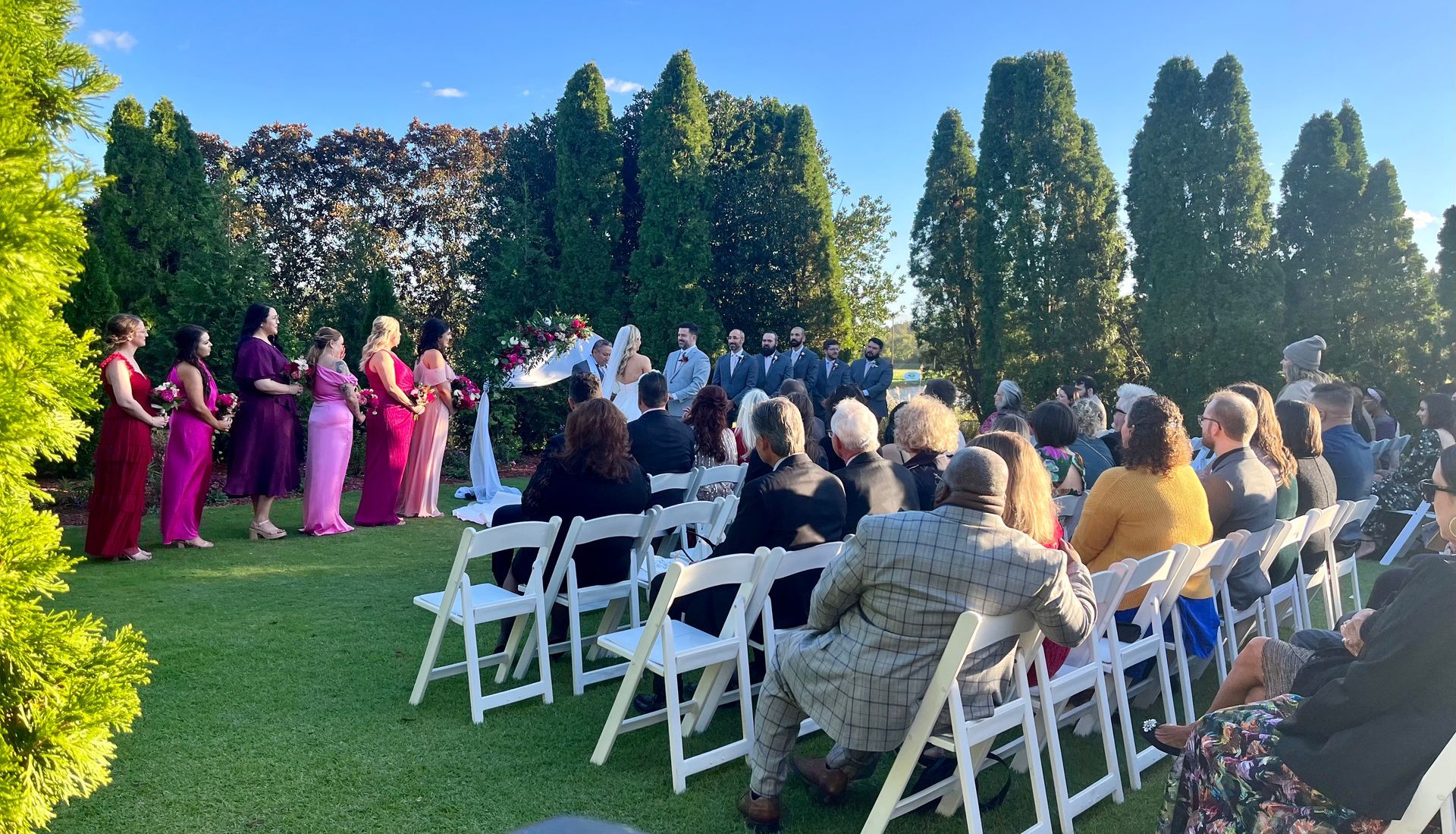 Outdoor wedding ceremony with guests seated in white chairs as attendants stand on a lawn near tall trees