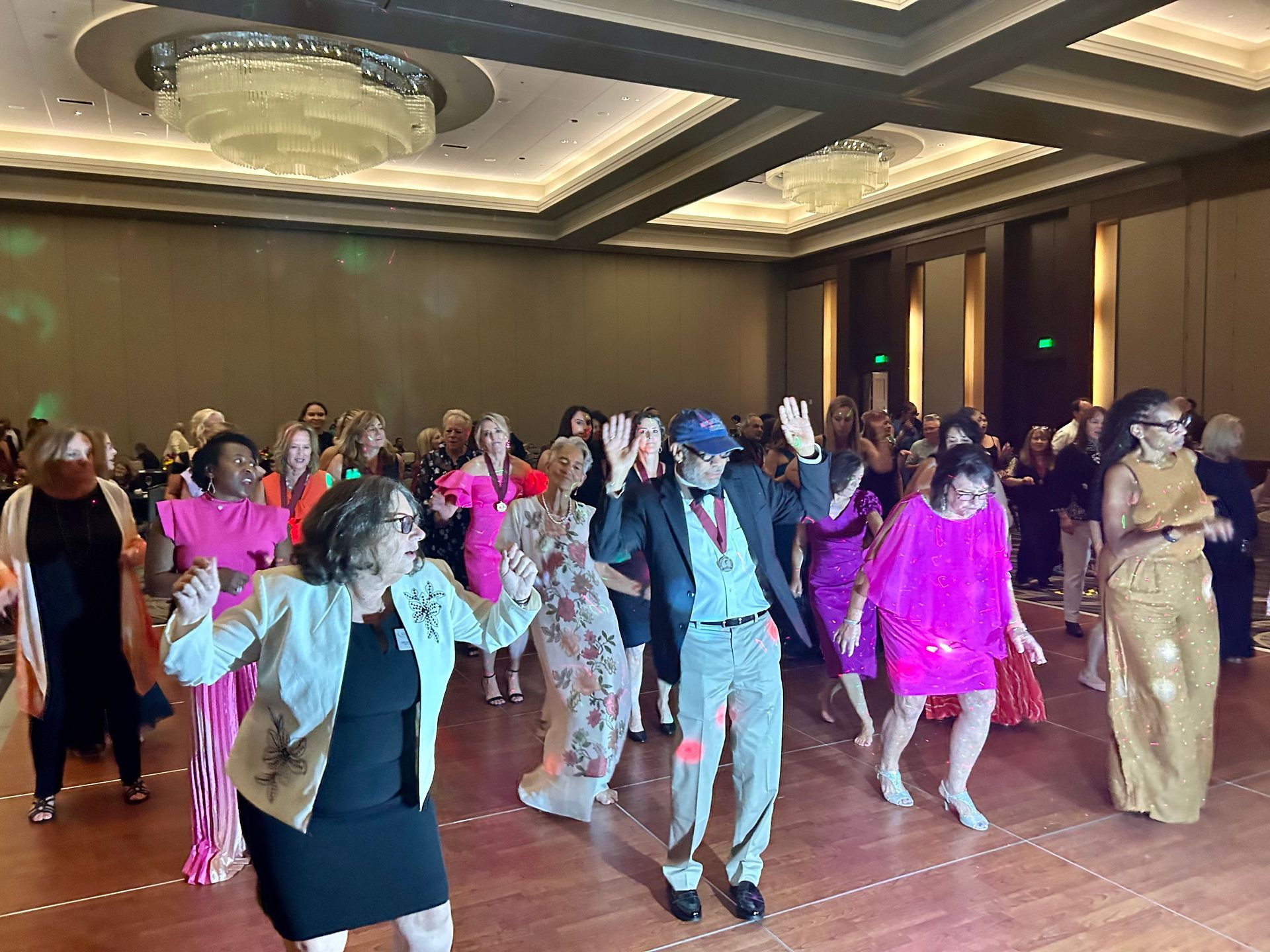People dancing in formal attire at a banquet hall, with bright pink and gold outfits under ceiling lights.