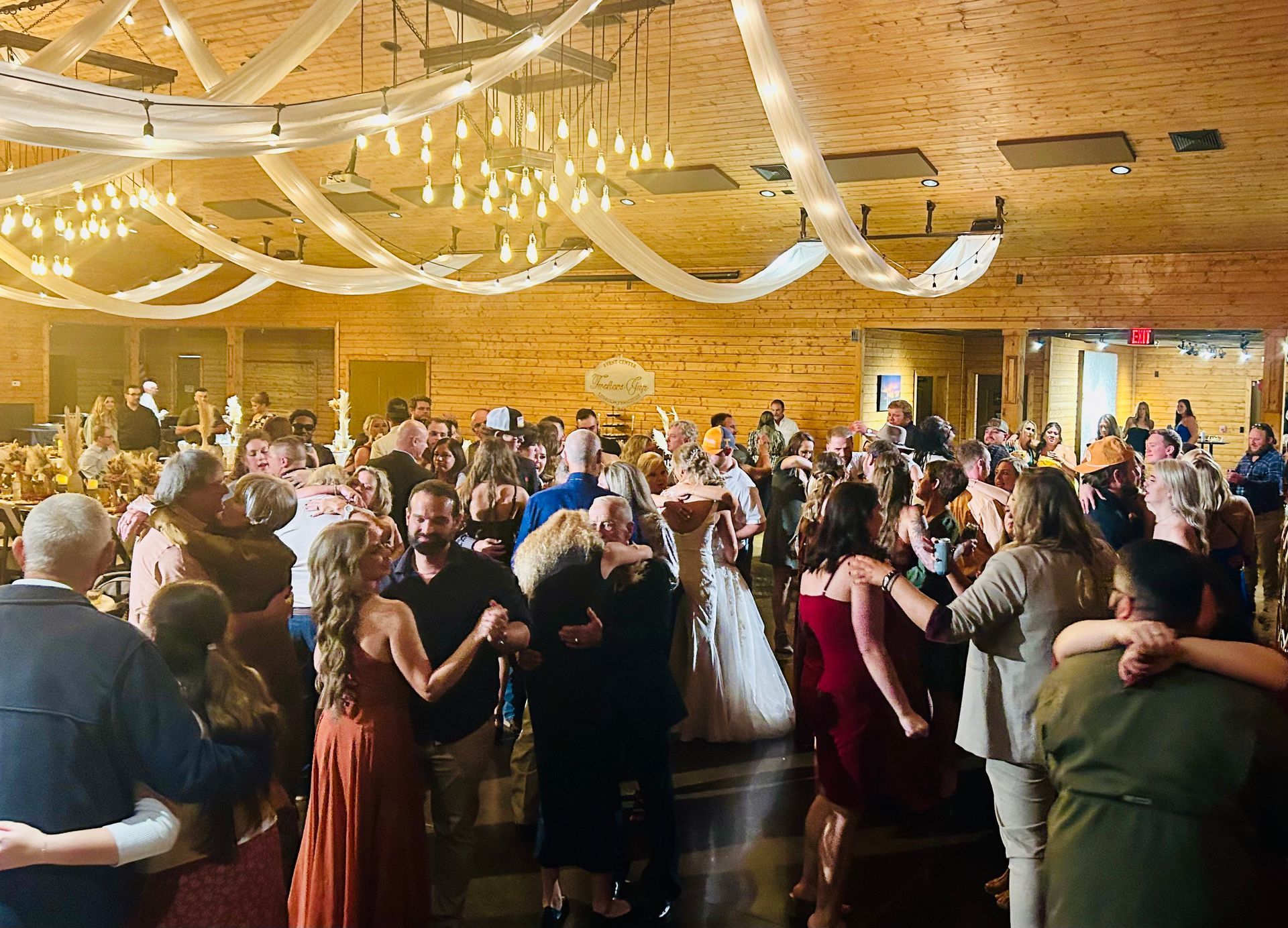 Crowded wedding reception in a wooden hall, with guests dancing under hanging white ribbons and warm lights
